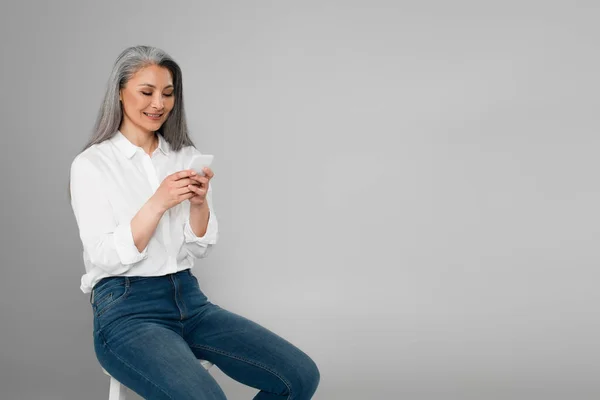 Feliz e elegante asiático mulher mensagens no smartphone enquanto sentado isolado no cinza — Fotografia de Stock