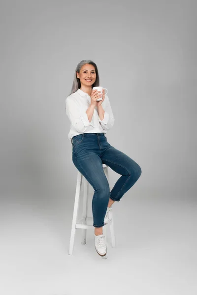 Happy asian woman in shirt and jeans sitting on high stool with tea on grey background — Stock Photo