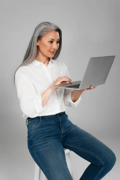 Mulher asiática de meia idade com cabelos grisalhos sentado nas fezes e digitando no laptop isolado no cinza — Fotografia de Stock
