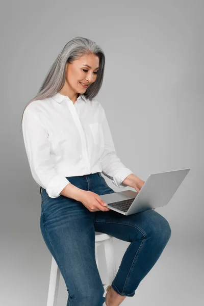Happy asian woman in white shirt typing on laptop while sitting on stool isolated on grey — Stock Photo