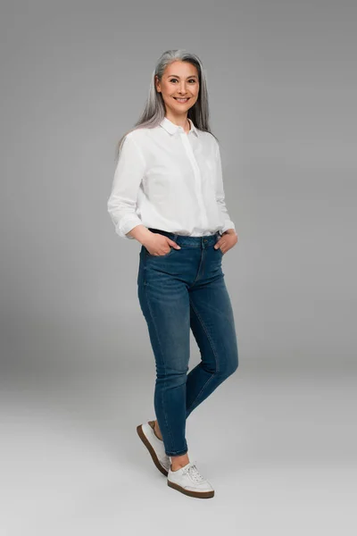 Vue pleine longueur de femme asiatique avec les mains dans des poches de jeans bleus souriant sur gris — Photo de stock