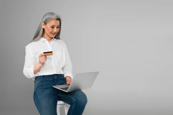 Усміхнена азіатська жінка з кредитною карткою сидить на табуреті і використовує ноутбук ізольовано на сірому — стокове фото