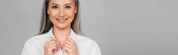 Mujer asiática feliz tocando cuello de camisa blanca aislado en gris, bandera - foto de stock