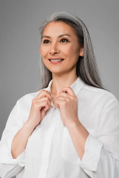 Зрелая азиатка улыбается, прикасаясь к воротнику белой рубашки, изолированной на сером — стоковое фото