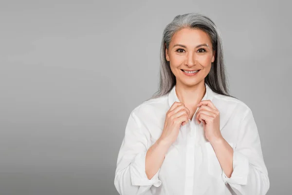 Pleased asian woman adjusting collar of white shirt while looking at camera isolated on grey — Stock Photo