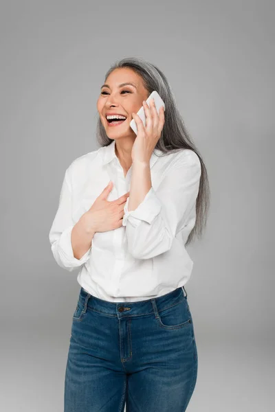 Mature asian woman touching chest and laughing while talking on mobile phone isolated on grey — Stock Photo