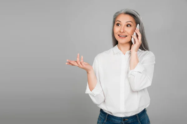 Excited asian woman in white shirt gesturing while talking on smartphone isolated on grey — Stock Photo