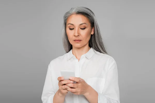 Serious asian woman messaging on mobile phone isolated on grey — Stock Photo