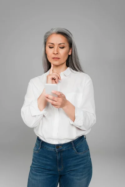 Thoughtful asian woman touching chin while looking at cellphone isolated on grey — Stock Photo