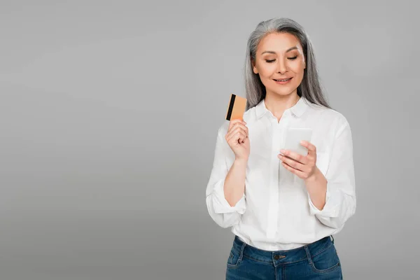 Smiling asian woman in white shirt holding credit card while looking at mobile phone isolated on grey — Stock Photo