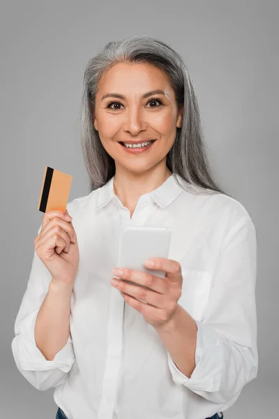 Stylish asian woman with credit card and mobile phone smiling at camera isolated on grey — Stock Photo