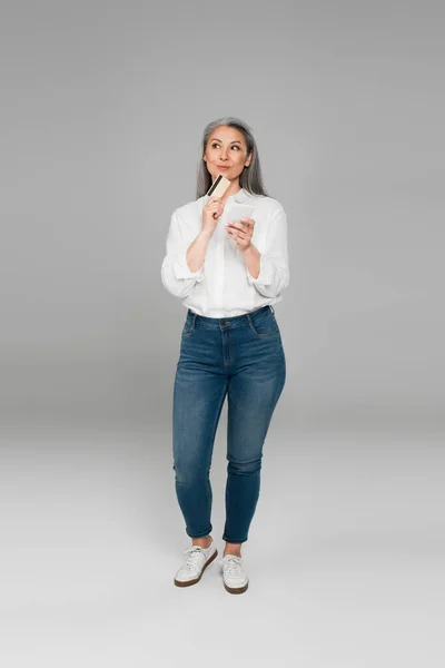 Sonriente y soñadora mujer asiática con tarjeta de crédito y teléfono móvil mirando hacia otro lado sobre fondo gris - foto de stock