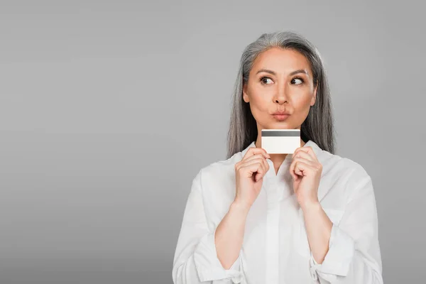 Thoughtful asian woman in white shirt holding credit card isolated on grey — Stock Photo