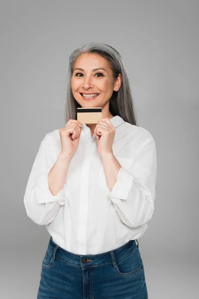 Задоволена азіатська жінка в білій сорочці, показуючи кредитну картку, дивлячись на камеру ізольовано на сірому — стокове фото