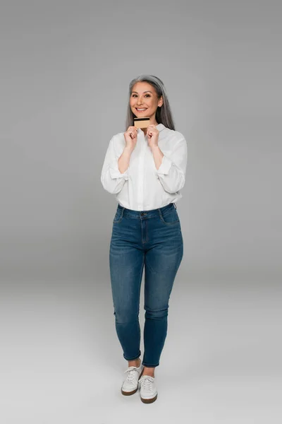 Longitud completa vista de sonriente asiático mujer mostrando tarjeta de crédito en gris fondo - foto de stock