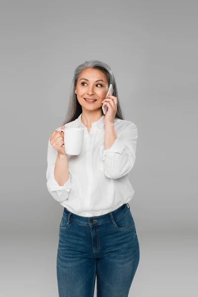 Positive asian woman holding cup of tea while talking on mobile phone isolated on grey — Stock Photo