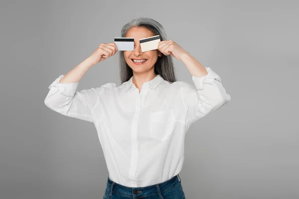 Alegre asiático mujer en blanco camisa cubriendo ojos con tarjetas de crédito aislado en gris — Stock Photo
