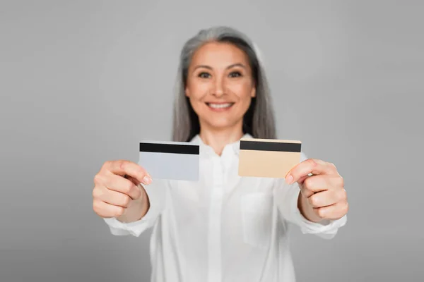 Verschwommene asiatische Frau lächelt, während sie Kreditkarten isoliert auf grau zeigt — Stockfoto
