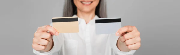 Обрізаний вид розмитої усміхненої жінки, що показує кредитні картки ізольовані на сірому, банер — стокове фото