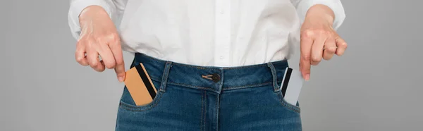 Vista parcial de la mujer que toma tarjetas de crédito de bolsillos de jeans aislados en gris, bandera - foto de stock