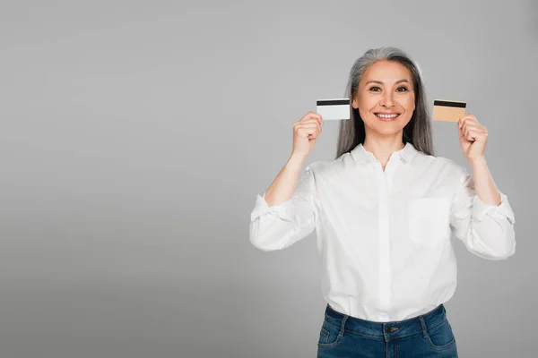 Радісна азіатська жінка в білій сорочці, що показує кредитні картки ізольовані на сірому — стокове фото