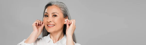 Lächelnde asiatische Frau, die Kopfhörer einsetzt, während sie die Kamera isoliert auf einem grauen Banner betrachtet — Stockfoto