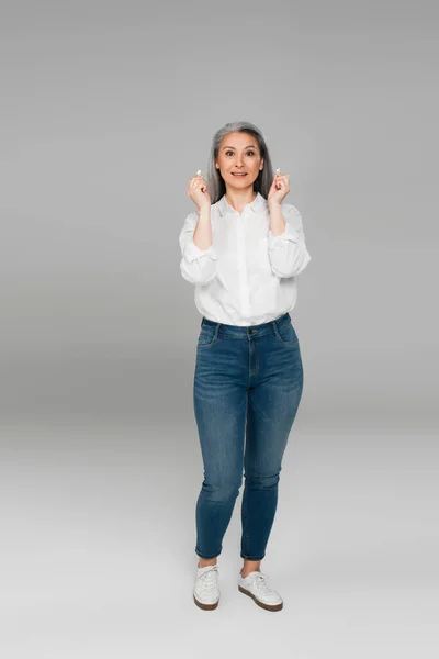 Повний вигляд зрілої азіатки в джинсах і сорочці, що тримає навушники на сірому — стокове фото