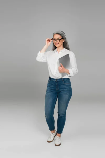 Mujer asiática positiva con portátil ajustando las gafas mientras mira hacia otro lado sobre fondo gris - foto de stock