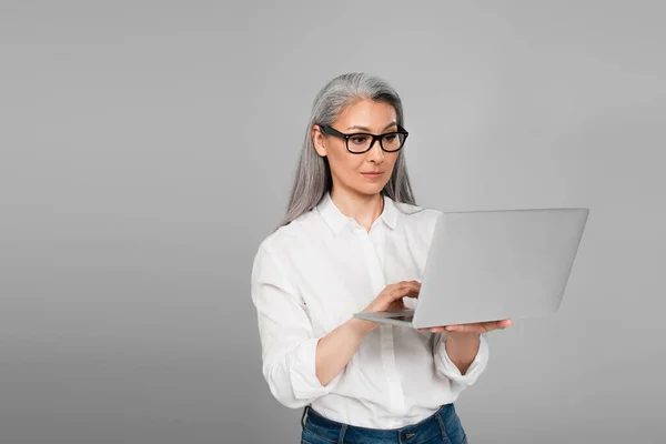 Mature asian woman in shirt and eyeglasses using laptop isolated on grey — Stock Photo