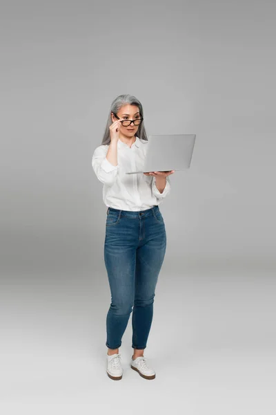 Full length view of asian woman adjusting eyeglasses while looking at laptop on grey background — Fotografia de Stock