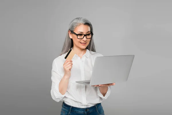 Freudige asiatische Frau schaut auf Laptop, während sie Kreditkarte isoliert auf grau hält — Stockfoto