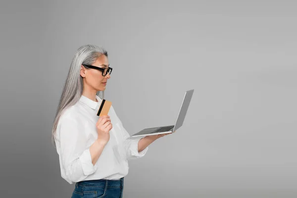Middle aged asian woman in white shirt and eyeglasses holding laptop and credit card isolated on grey — Stock Photo