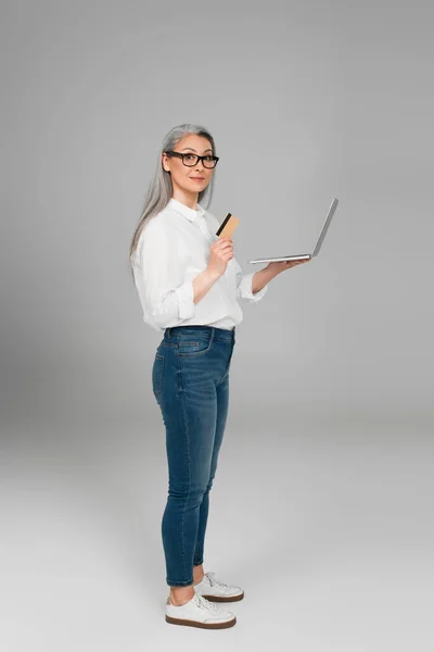 Mujer asiática madura en jeans y camisa blanca de pie con tarjeta de crédito y portátil sobre fondo gris - foto de stock