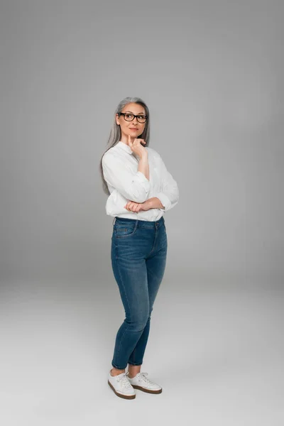 Full length view of mature asian woman in jeans, white shirt and eyeglasses standing on grey — Fotografia de Stock