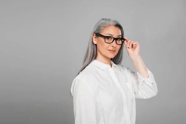 Middle aged asian woman in white shirt looking at camera while touching eyeglasses isolated on grey — Stock Photo