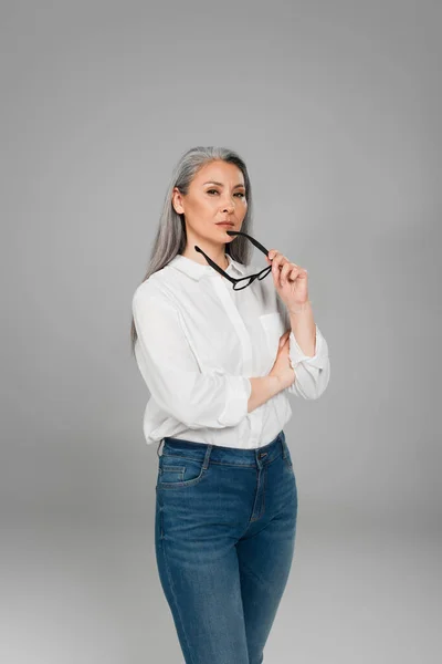 Confident asian woman in jeans and white shirt holding eyeglasses isolated on grey — Stock Photo