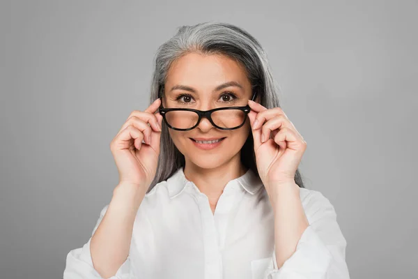 Зріла азіатська жінка в білій сорочці знімає окуляри, посміхаючись на камеру ізольовано на сірому — стокове фото