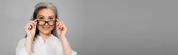 Mujer asiática positiva sonriendo a la cámara mientras se quita las gafas aisladas en gris, pancarta - foto de stock