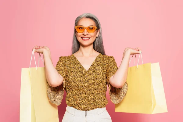 Alegre asiático mujer en elegante ropa celebración compras en rosa fondo - foto de stock