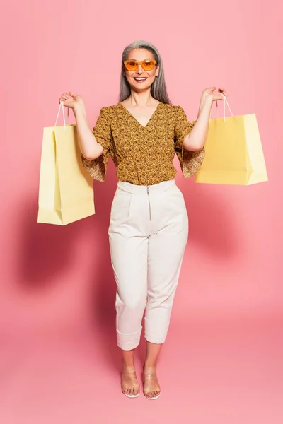 Full length view of mature asian woman in trendy clothes holding shopping bags on pink background — Stock Photo