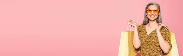 Felice donna asiatica in occhiali da sole gialli e camicetta fantasia in piedi con borse della spesa isolate su rosa, banner — Foto stock