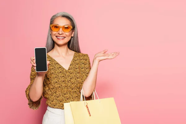 Mujer asiática con estilo con bolsa de compras y teléfono inteligente con pantalla en blanco aislado en rosa - foto de stock