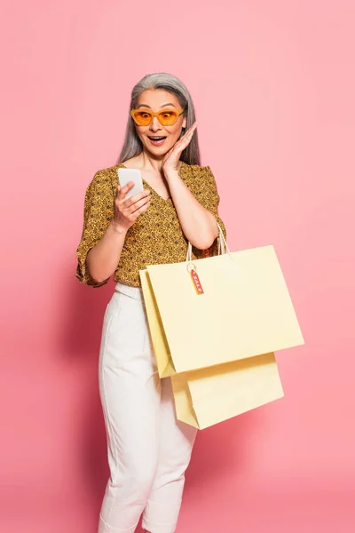 Mujer asiática asombrada con bolsas de compras amarillas mirando el teléfono celular sobre fondo rosa - foto de stock