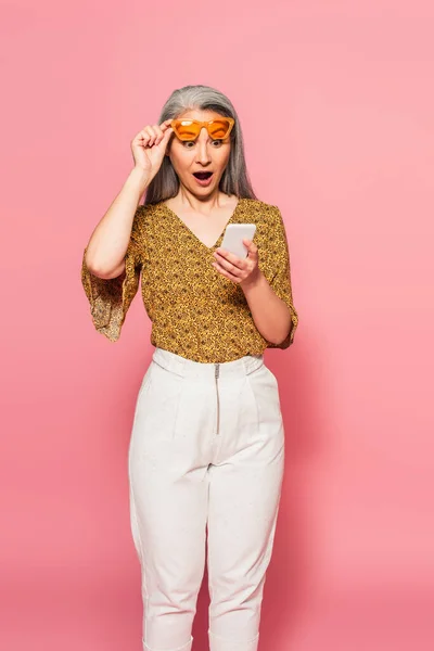 Erstaunt asiatische Frau nimmt Sonnenbrille ab, während sie auf Handy auf rosa Hintergrund schaut — Stockfoto