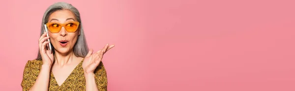 Thrilled asian woman pointing with hand while talking on cellphone isolated on pink, banner — Stock Photo