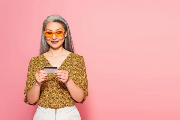 Mature asian woman in yellow sunglasses looking at credit card isolated on pink — Stock Photo