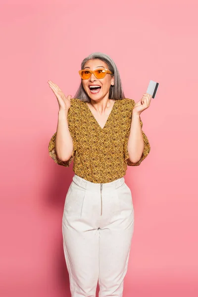 Sobregocijado asiático mujer con tarjeta de crédito mostrando wow gesto en rosa fondo - foto de stock