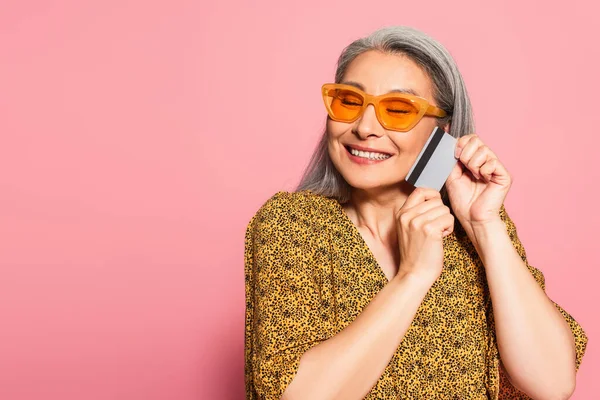Mujer asiática feliz en gafas de sol de moda celebración de la tarjeta de crédito cerca de la cara aislado en rosa - foto de stock