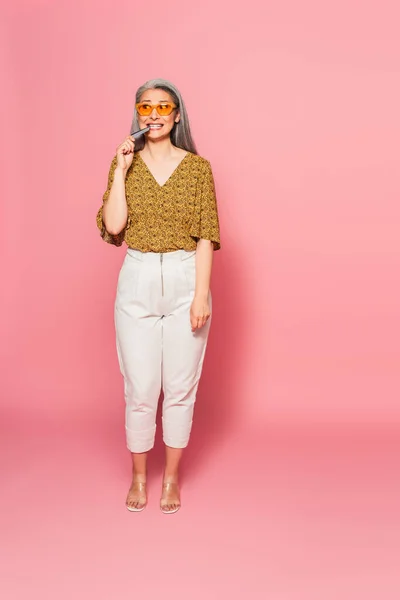 Mujer asiática nerviosa en ropa de moda mordiendo tarjeta de crédito mientras mira hacia otro lado en el fondo rosa - foto de stock