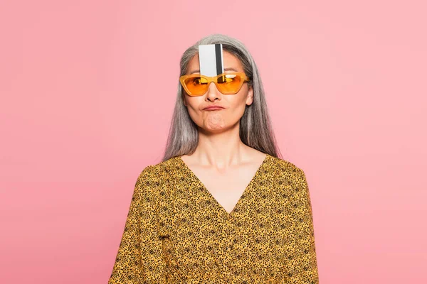 Mature asiatique femme avec carte de crédit entre lunettes de soleil et front grimacing isolé sur rose — Photo de stock